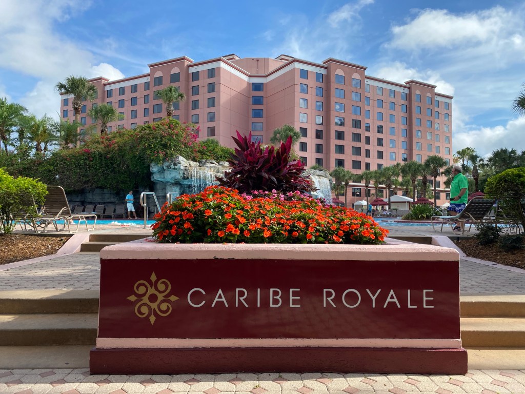 A sign with the words "Caribe Royale" is in the foreground with red impatien flowers just before two steps that lead up to a pool deck. The pool with a waterful is in the middle ground and the hotel building is soft pink in the background beneath blue skits.